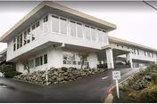 Exterior view of a two-story white building with large windows and a covered entrance. There is a paved driveway with a sign that reads 'EXIT ONLY' and landscaping with rocks and plants around the building.