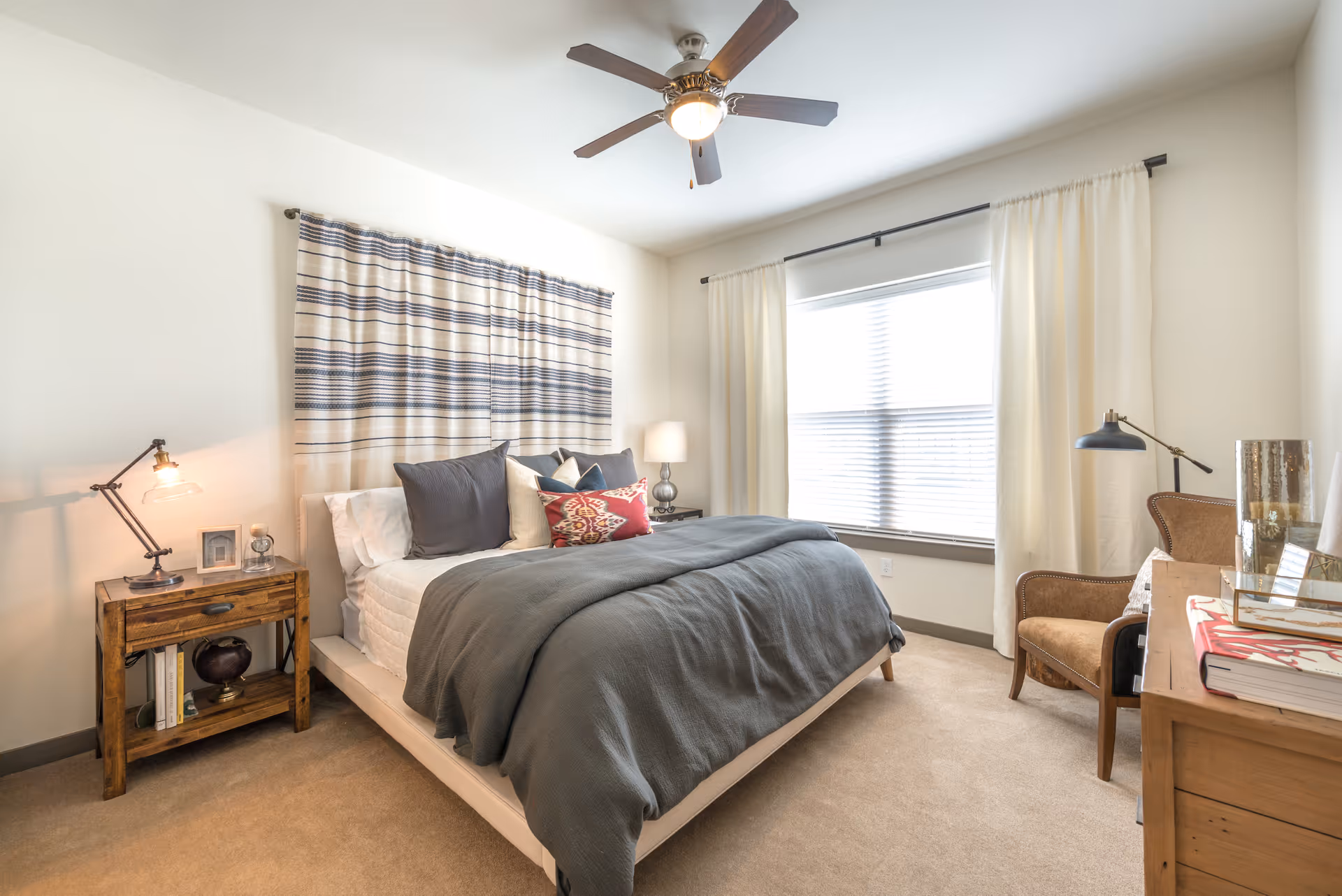 A bright bedroom with a large bed covered in gray bedding and multiple pillows, a wooden nightstand with a lamp and decorative items, a window with white curtains letting in natural light, a ceiling fan with a light, and a wooden dresser with a chair and desk lamp beside it.