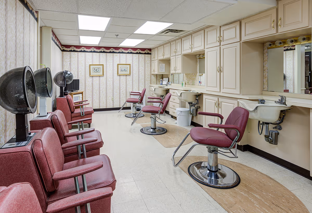 Interior of a hair salon with multiple maroon salon chairs, hair dryers, sinks, and mirrors along the walls. The room has patterned wallpaper, overhead fluorescent lighting, and cabinetry for storage.