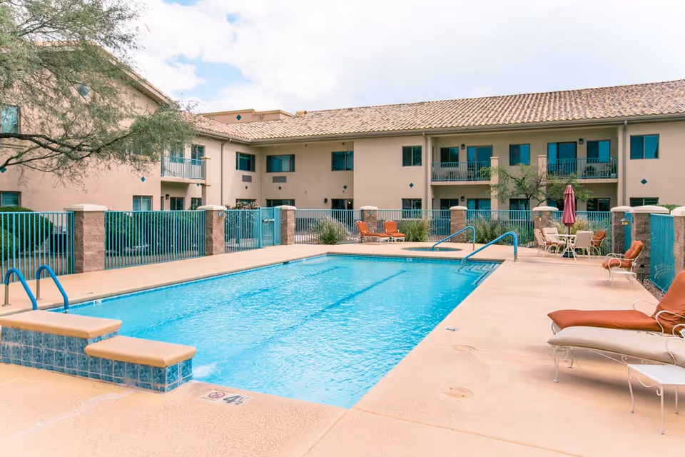 Outdoor swimming pool surrounded by lounge chairs and a fenced patio with a two-story beige apartment building in the background.
