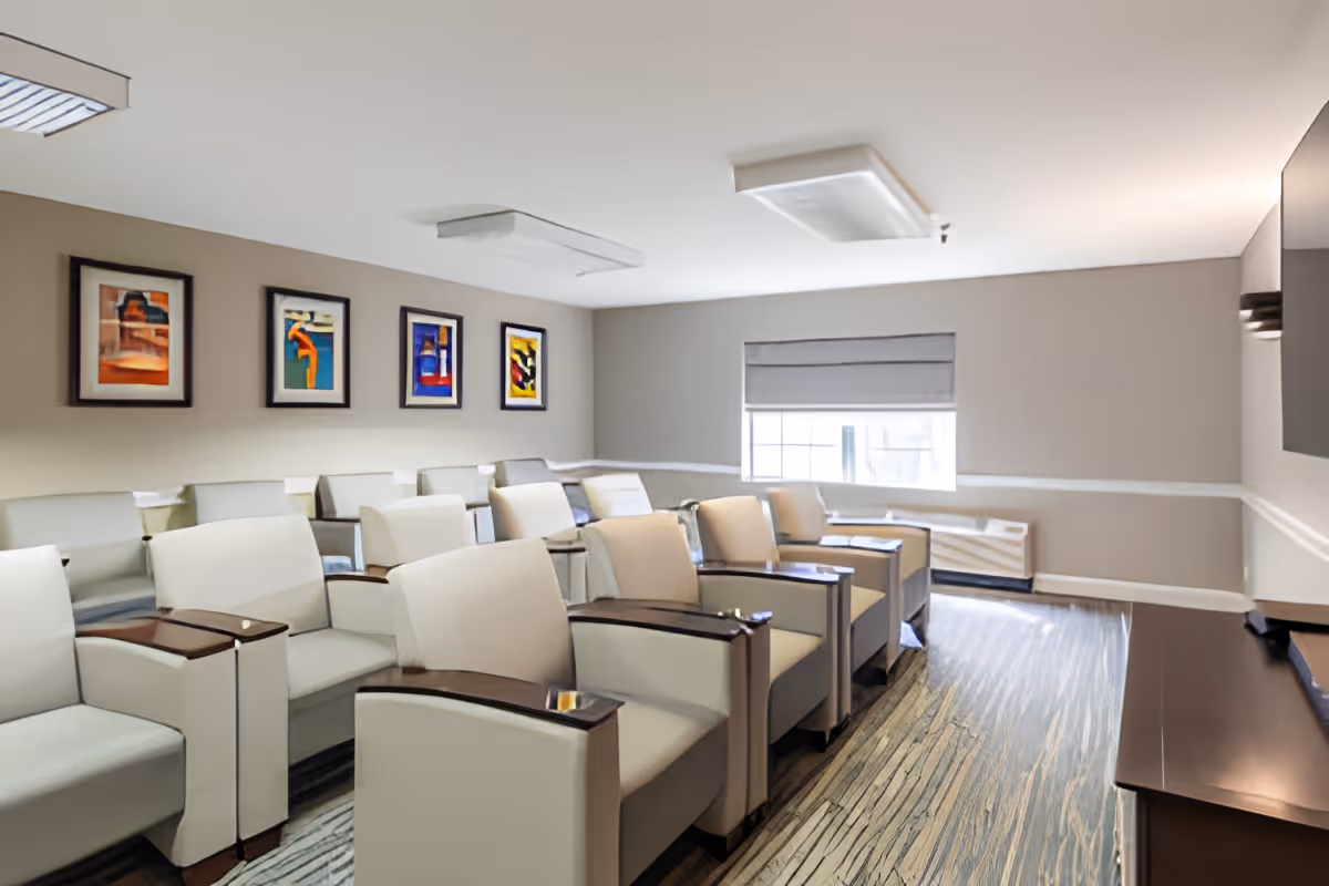 A small theater or media room with several rows of beige armchairs with wooden armrests facing a large flat-screen TV mounted on the wall. The room has neutral-colored walls, a window with a gray roller shade, and four colorful framed artworks hanging on one wall. The floor has a patterned carpet, and the ceiling has rectangular light fixtures.