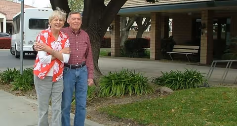 An elderly couple standing outside near a tree and garden area in front of a building with a covered porch and benches. The woman is wearing a red floral shirt and light pants, and the man is wearing a red checkered shirt and blue jeans.