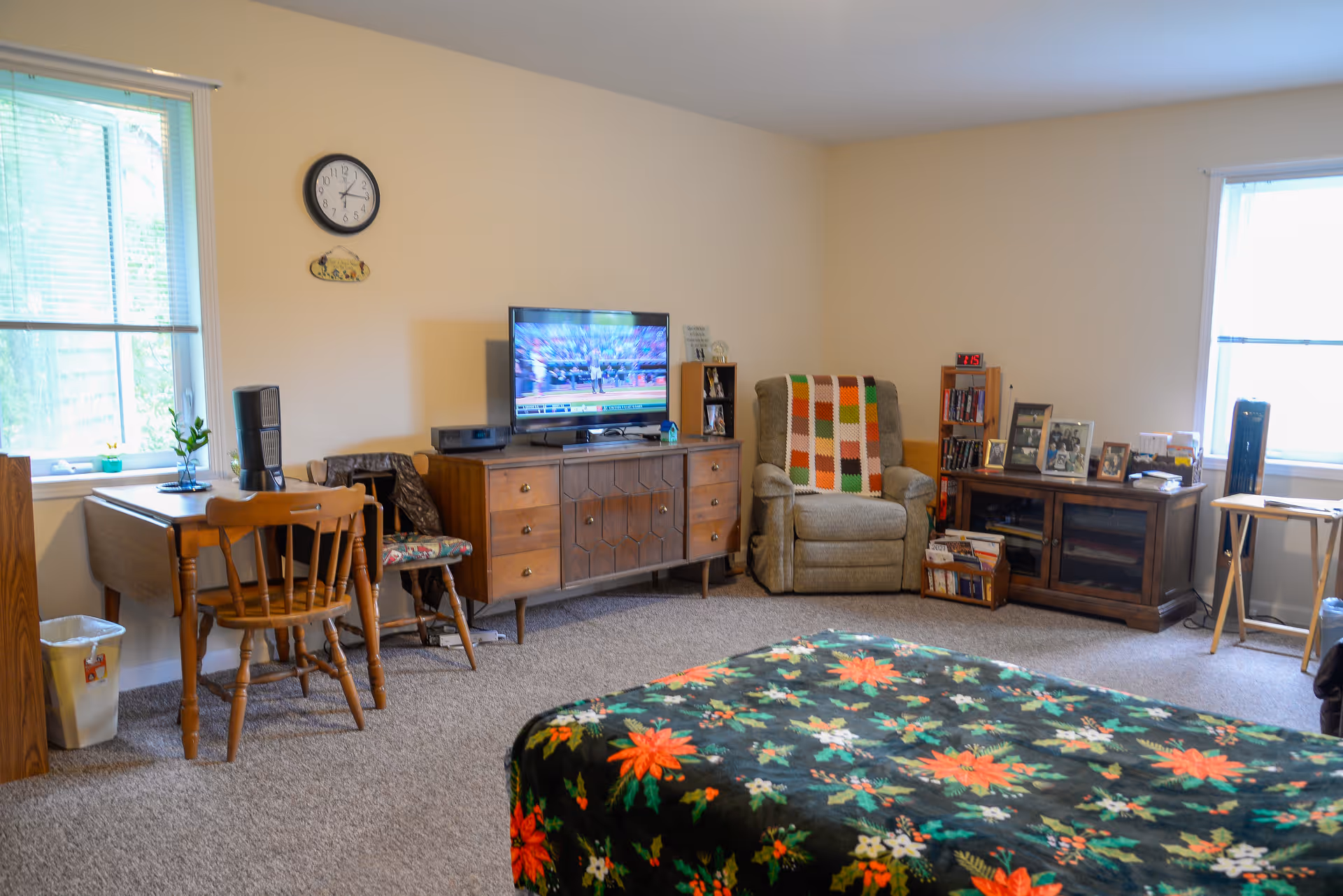 A cozy living space with a floral patterned bedspread in the foreground, a wooden desk and chair near a window on the left, a wooden dresser with a flat-screen TV showing a sports game, a comfortable armchair with a colorful crocheted blanket, and a wooden cabinet with framed photos and books. The room has beige walls and carpeted floor with two windows letting in natural light.