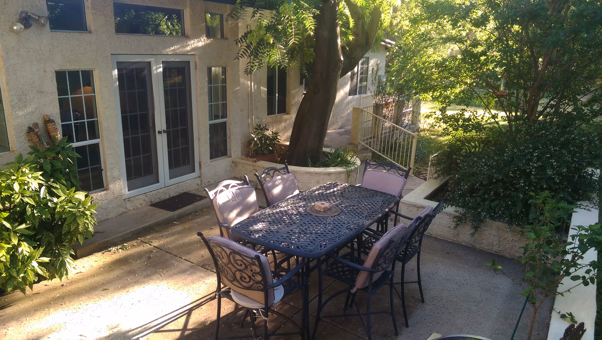 Outdoor patio area with a black metal table and six cushioned chairs surrounded by greenery and trees, adjacent to a building with glass doors and windows.