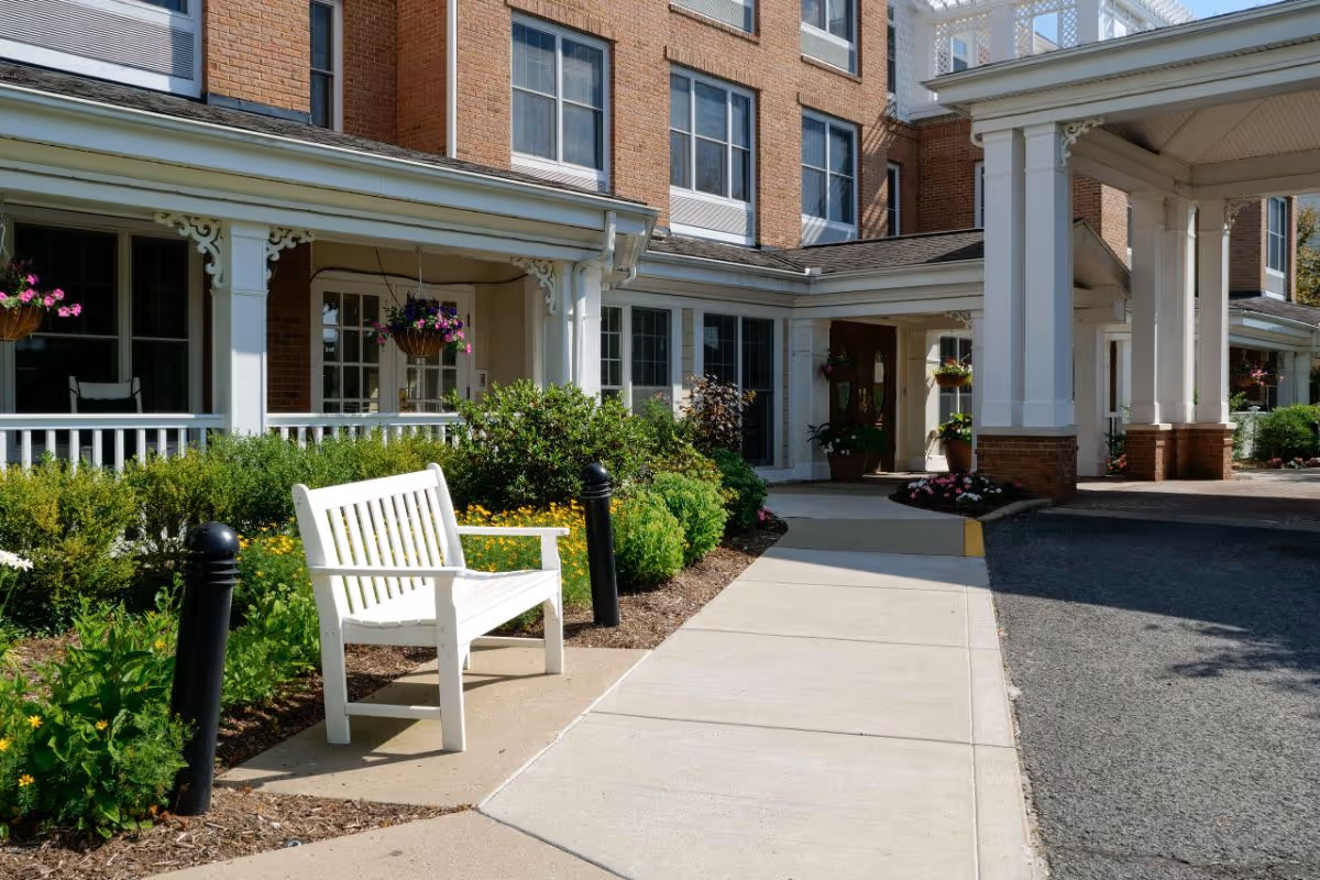 Exterior view of a senior living facility showing a white bench on a sidewalk surrounded by green bushes and flowers, with a brick building and covered entrance in the background.