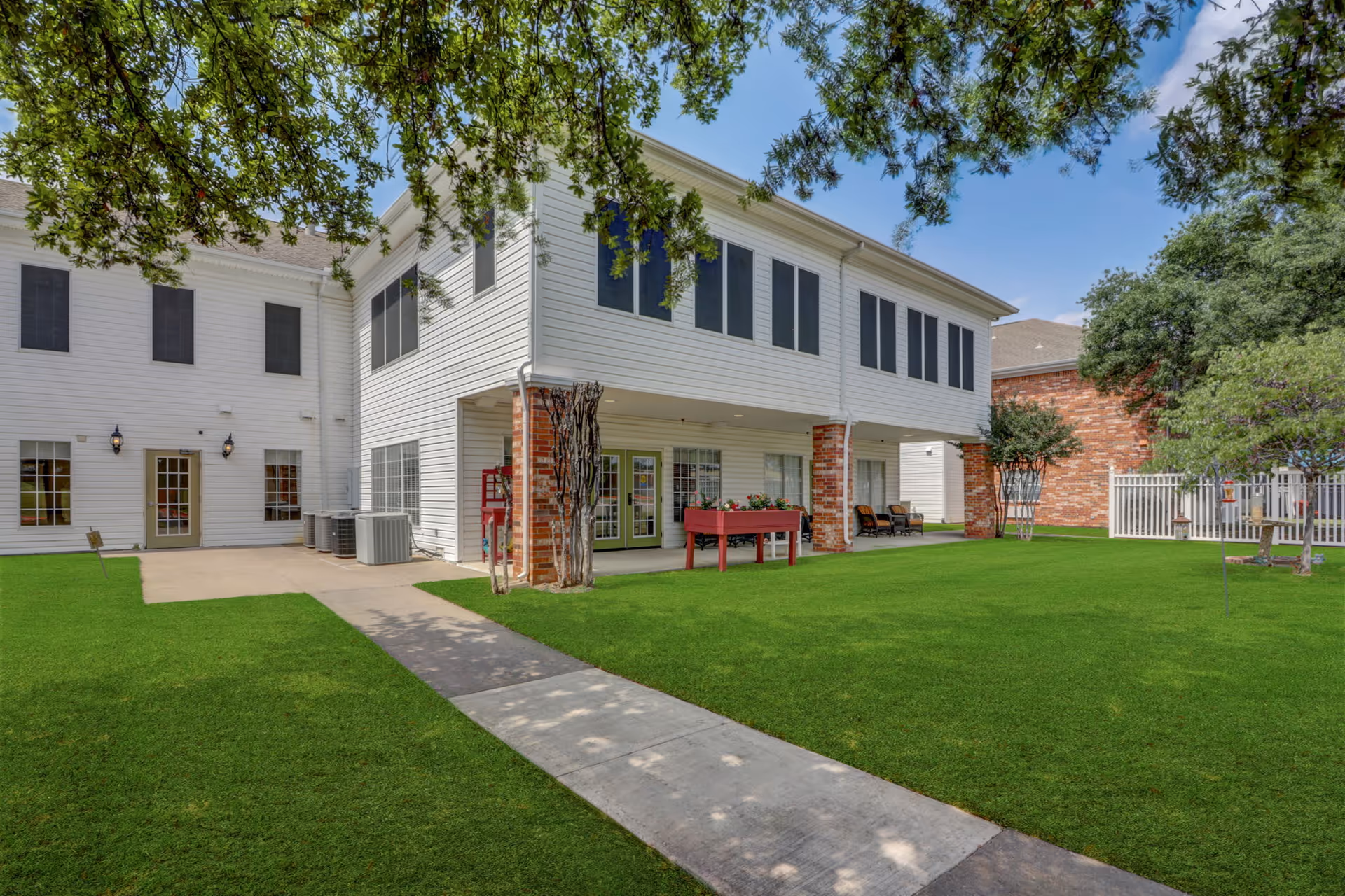 Exterior view of a senior living facility building with white siding and brick pillars. The building has multiple windows and a covered patio area with chairs and a raised garden bed. There is a concrete walkway leading through a well-maintained green lawn with trees providing shade.