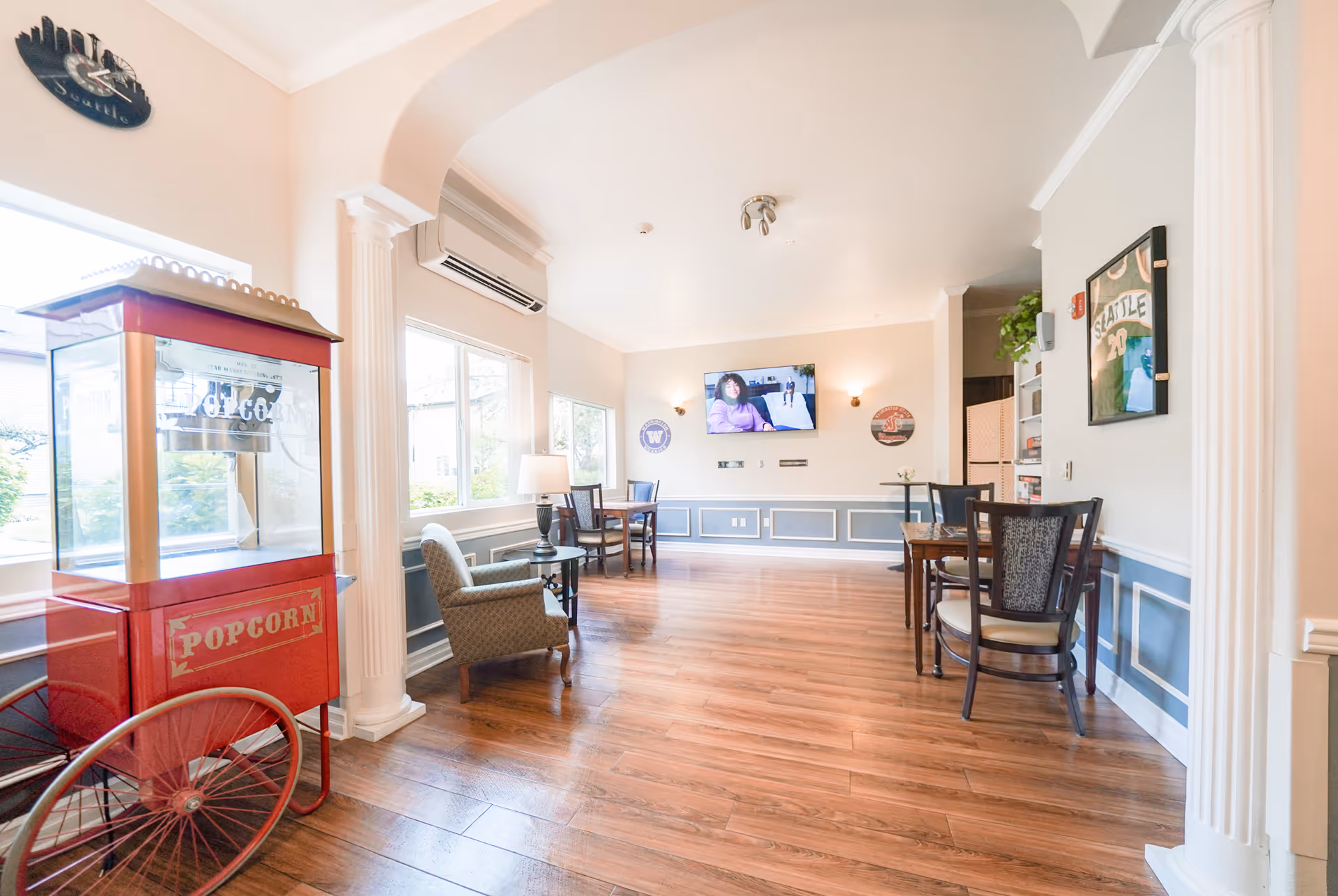 A bright and spacious senior living common area with wooden floors, white walls, and large windows letting in natural light. The room features a red vintage-style popcorn machine on the left, a comfortable armchair, several tables and chairs, decorative columns, and a wall-mounted TV displaying a woman. Various wall decorations and a clock are also visible.