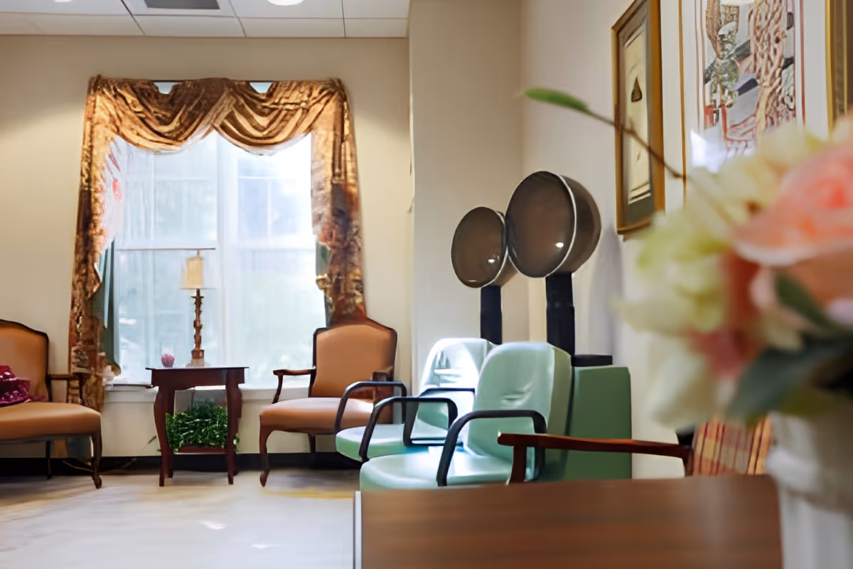 A cozy room with two vintage hair dryer chairs in light green, two beige armchairs, a small wooden table with a lamp, and a large window with ornate brown curtains. There are framed artworks on the wall and a blurred bouquet of flowers in the foreground.
