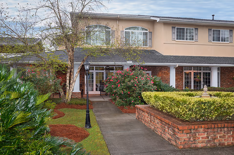 Exterior view of The Bridge at Charleston facility showing a two-story building with beige and brick walls, large windows, a walkway leading to the entrance, surrounded by well-maintained landscaping including bushes, flowering plants, and a tree.