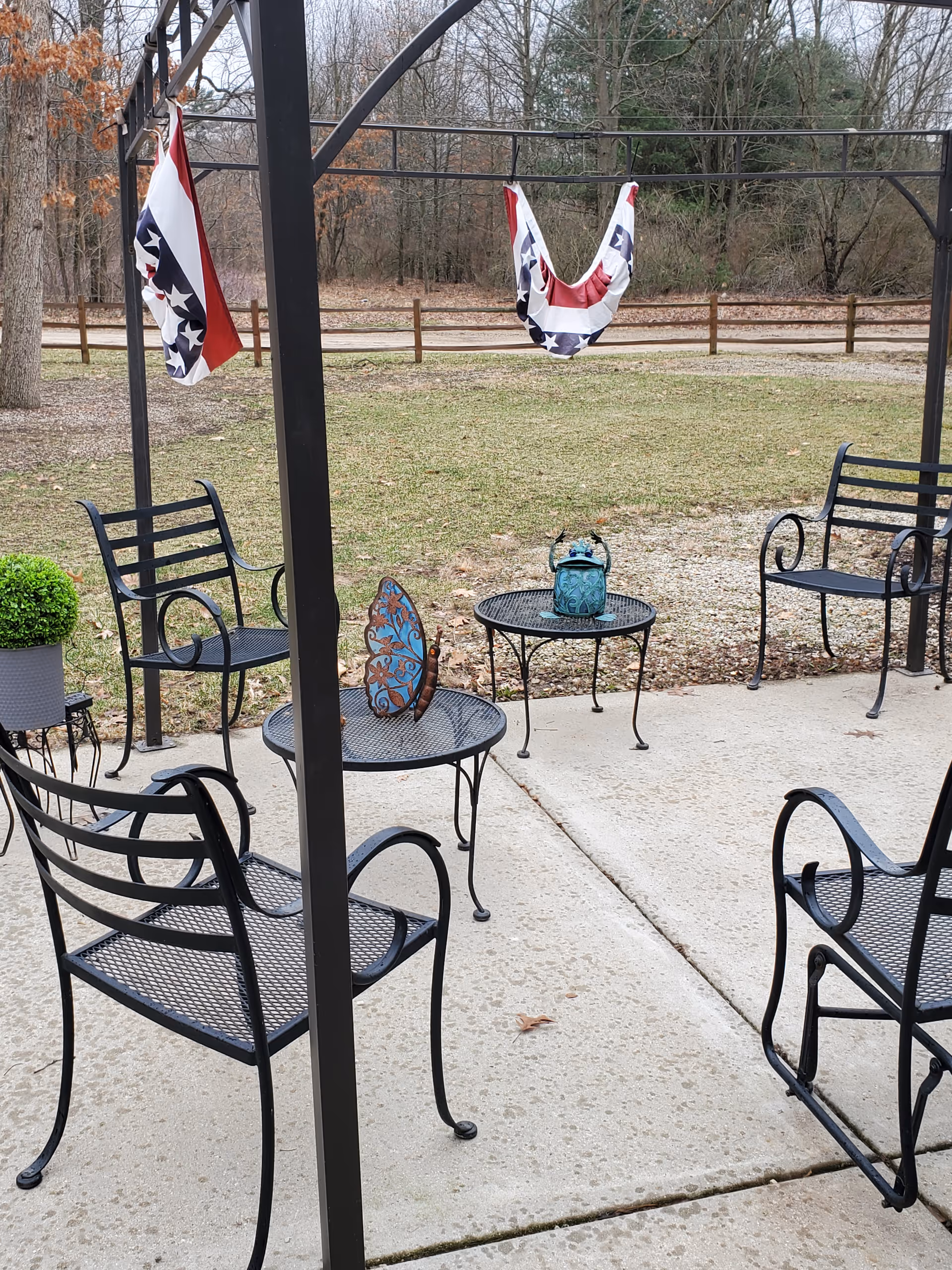 Outdoor patio area with black metal chairs and tables on a concrete surface. Two patriotic red, white, and blue fabric decorations hang from a metal frame. A small decorative blue butterfly and a teal lantern are placed on the tables. In the background, there is a grassy area with a wooden fence and leafless trees.