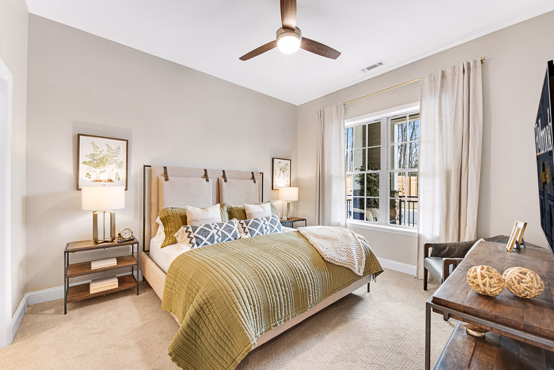 A bright and cozy bedroom with a large bed featuring a beige upholstered headboard, green quilted bedspread, and multiple pillows. There are two bedside tables with lamps and framed botanical prints above them. A ceiling fan with a light is mounted on the white ceiling. A large window with beige curtains lets in natural light, and a wooden dresser with decorative items and a TV is positioned opposite the bed.