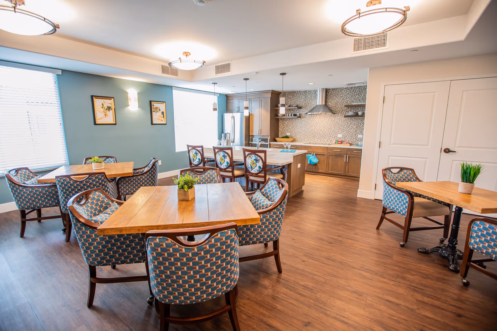 Bright communal dining area with wooden tables, patterned chairs, and an open kitchen in the background.