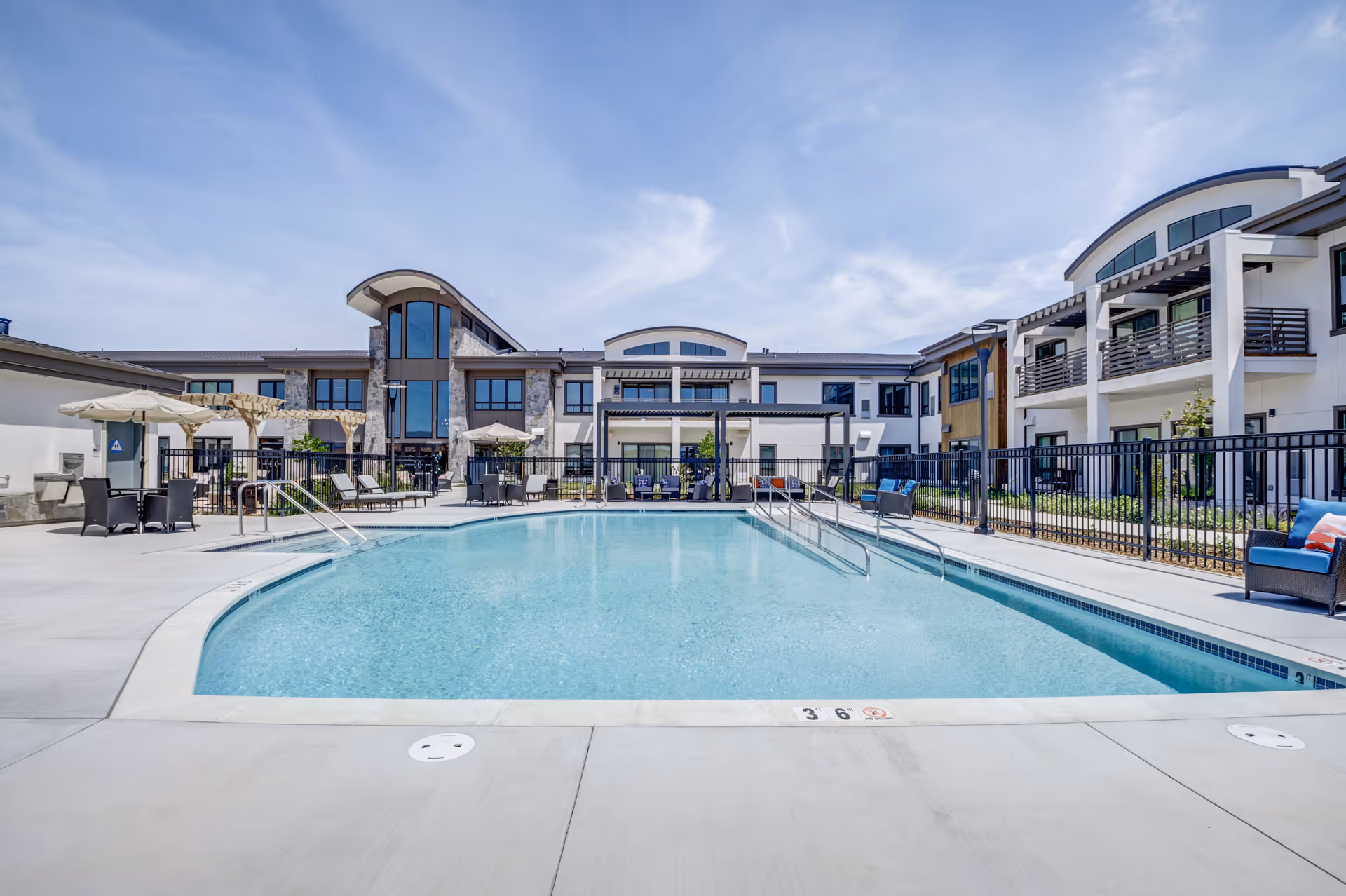 Outdoor swimming pool with clear blue water surrounded by a concrete deck, lounge chairs, tables with umbrellas, and a modern two-story building in the background under a partly cloudy sky.