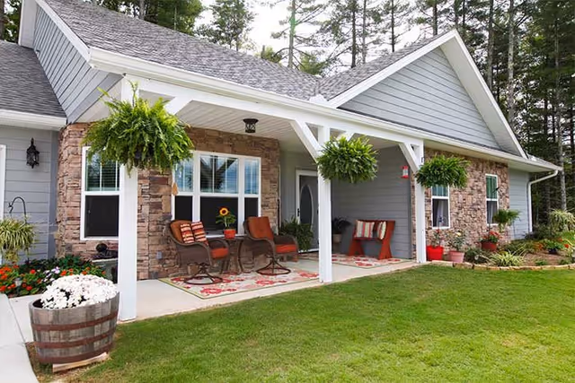 Front porch of a single-story house with stone and gray siding exterior, featuring two hanging ferns, two brown chairs with red cushions, a small table with a sunflower in a pot, a red bench with striped pillows, and various potted plants around the porch. The house is surrounded by green grass and trees in the background.