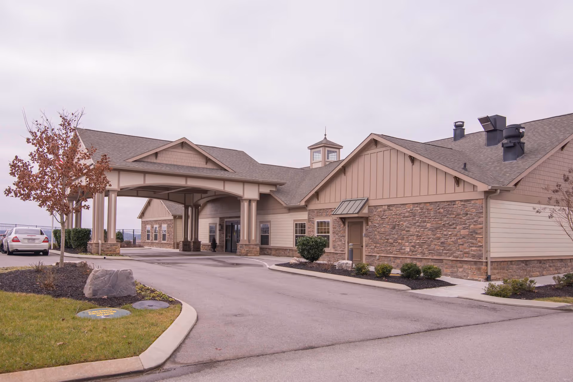 Exterior view of Morning Pointe of Lenoir City facility showing a covered entrance with stone and beige siding, a driveway, landscaped bushes, a tree with brown leaves, and a parked white car.