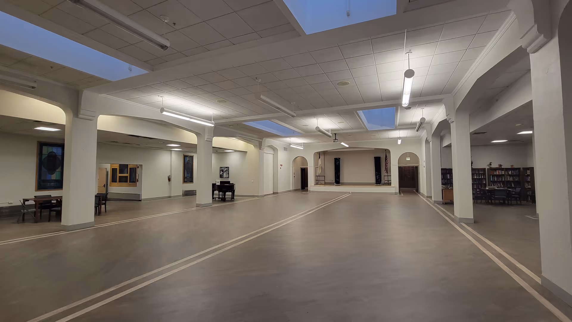 A large, open interior space with a stage at the far end, several tables and chairs along the sides, and bookshelves visible on the right. The ceiling has multiple skylights and hanging lights, and the floor is smooth with light-colored lines marking sections.