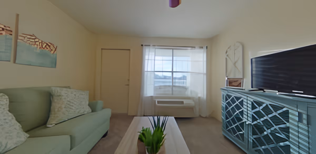 A living room with a green sofa on the left, a white coffee table with a small green plant in the center, and a blue TV stand with a flat-screen television on the right. There is a window with sheer white curtains and a door next to it at the back of the room. The walls are light-colored and there are two abstract paintings above the sofa.