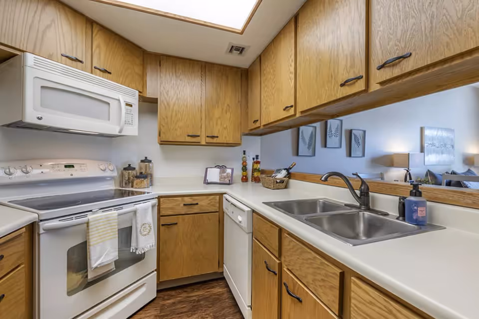 A kitchen with wooden cabinets, a white electric stove with two towels hanging on the handle, a white microwave above the stove, a double stainless steel sink with a faucet, a dishwasher, and a countertop with various items including a soap dispenser and decorative bottles. A large mirror above the sink reflects part of a living room with wall art and lamps.