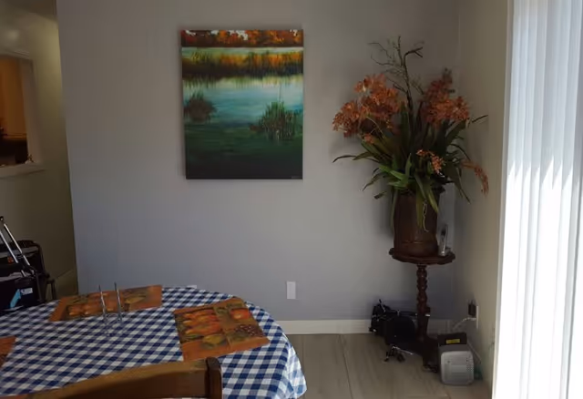 A dining area with a round table covered with a blue and white checkered tablecloth and four orange placemats. On the wall above the table is a painting depicting a serene water scene with greenery and autumn-colored trees. To the right, there is a small wooden side table with a large vase holding orange flowers and green leaves. Vertical blinds cover a window or door on the right side, allowing natural light to enter the room.