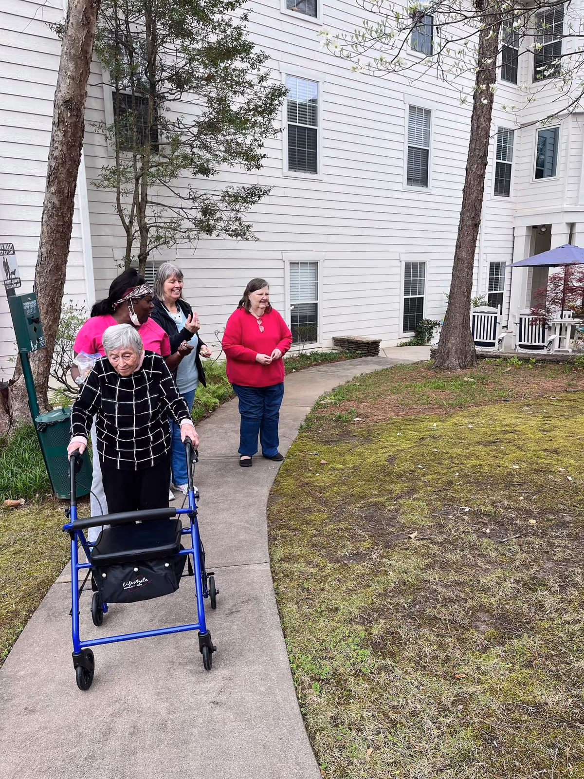 An elderly woman using a blue walker is walking on a paved path outside a white multi-story building. Behind her, three women stand on the path, smiling and watching her. The area has some trees, grass, and a small patio with a purple umbrella and white chairs.