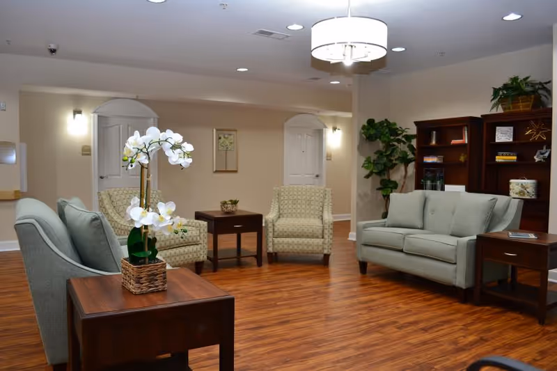 A cozy living room area with wooden flooring, light green upholstered chairs and sofa, wooden side tables, a bookshelf with decorative items, and a potted plant. The room is softly lit with ceiling lights and wall sconces, and there are two white doors in the background.