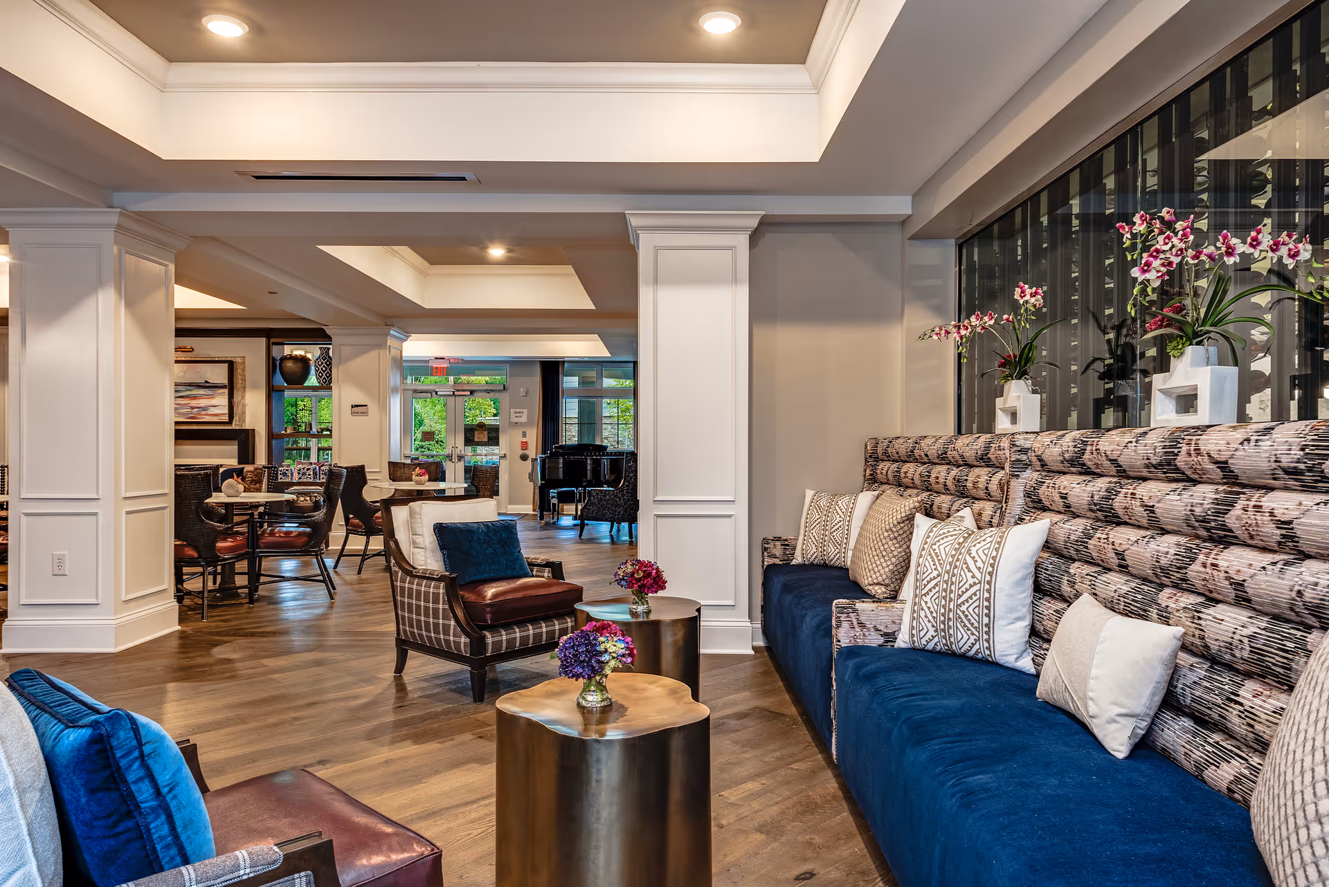 A spacious and elegantly decorated common area in a senior living facility featuring a long cushioned bench with patterned pillows and vases of flowers on a glass shelf above it. There are also comfortable armchairs with blue and brown cushions, small round tables with flower arrangements, and a grand piano in the background near large windows and glass doors letting in natural light.