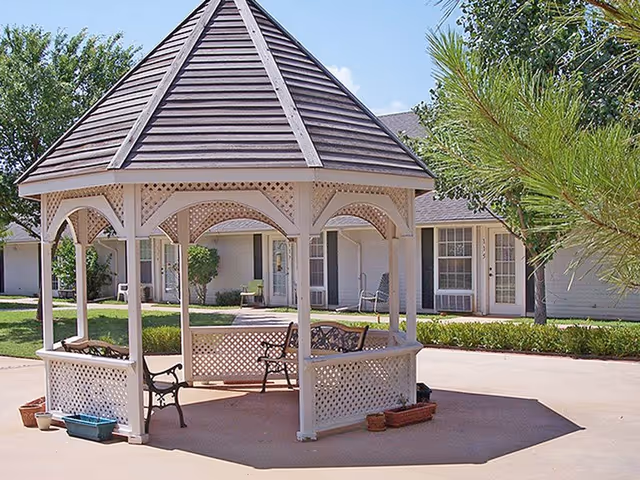 A white wooden gazebo with lattice panels and benches inside, situated on a concrete patio area with green grass, trees, and a senior living facility building in the background.