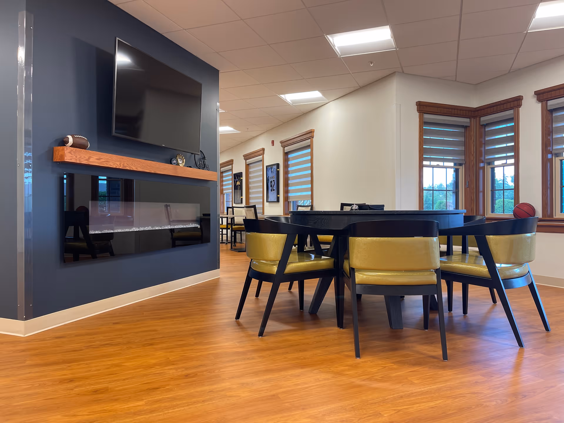Bright communal lounge with a round table surrounded by yellow-upholstered chairs, a wall-mounted TV and modern fireplace, and windows along the right wall.