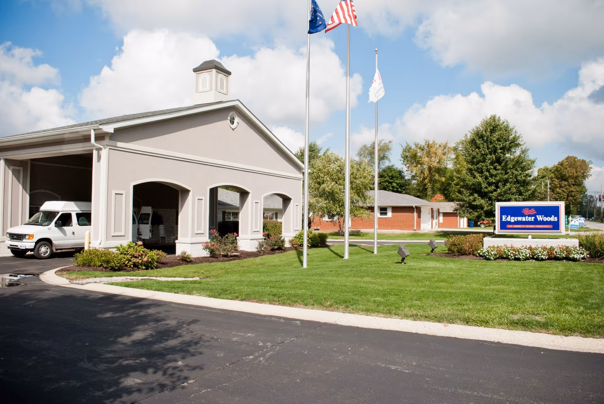Exterior view of Edgewater Woods senior living facility showing a covered entrance with a white van parked underneath, three flagpoles with flags, a well-maintained lawn, and a sign that reads 'Edgewater Woods An American Senior Community'.