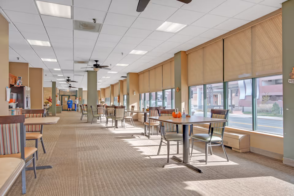 A spacious dining area with multiple tables and chairs arranged along large windows with beige blinds. The room has a carpeted floor, ceiling fans, and overhead lighting. Two people are visible in the background near a kitchen or service area.