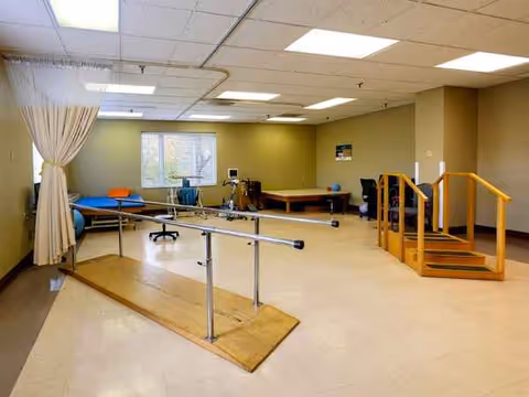 Rehabilitation therapy room with parallel bars, stairs, mats, and exercise equipment.