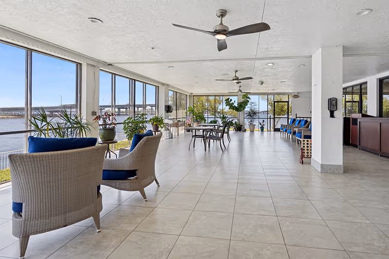 A spacious indoor lounge area with large windows offering a view of a waterfront and a bridge. The room features tiled floors, ceiling fans, wicker chairs with blue cushions, tables with chairs, potted plants, and a row of blue cushioned chairs along one wall.