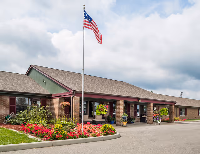 Exterior view of a single-story brick building with a pitched roof, an American flag on a flagpole, and colorful flower beds in front. The building has a covered entrance with seating and hanging flower baskets.