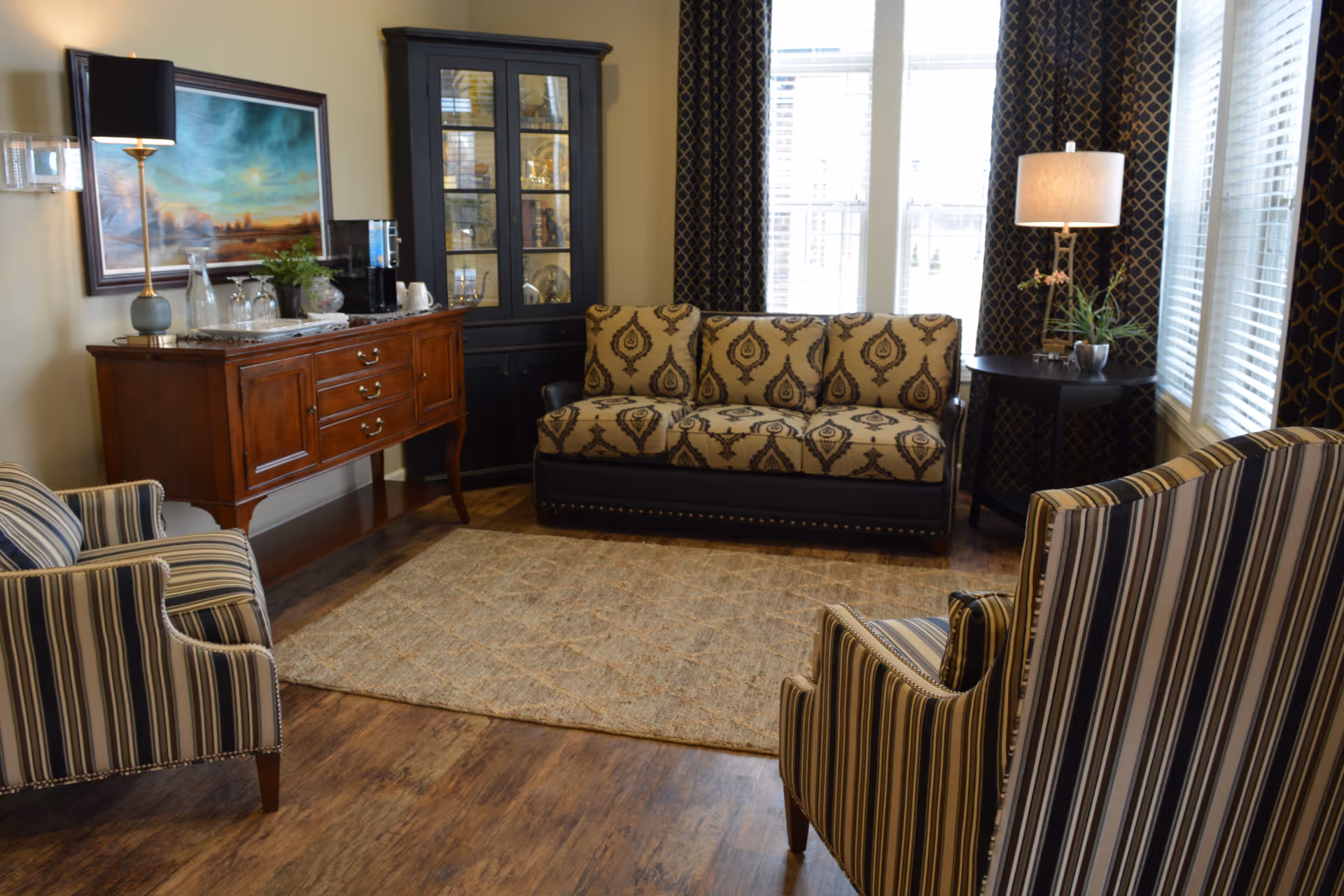 A cozy living room with a patterned sofa, two striped armchairs, a wooden sideboard with a lamp and decorative items, a black display cabinet, a round side table with a lamp and plant, and large windows with patterned curtains letting in natural light.