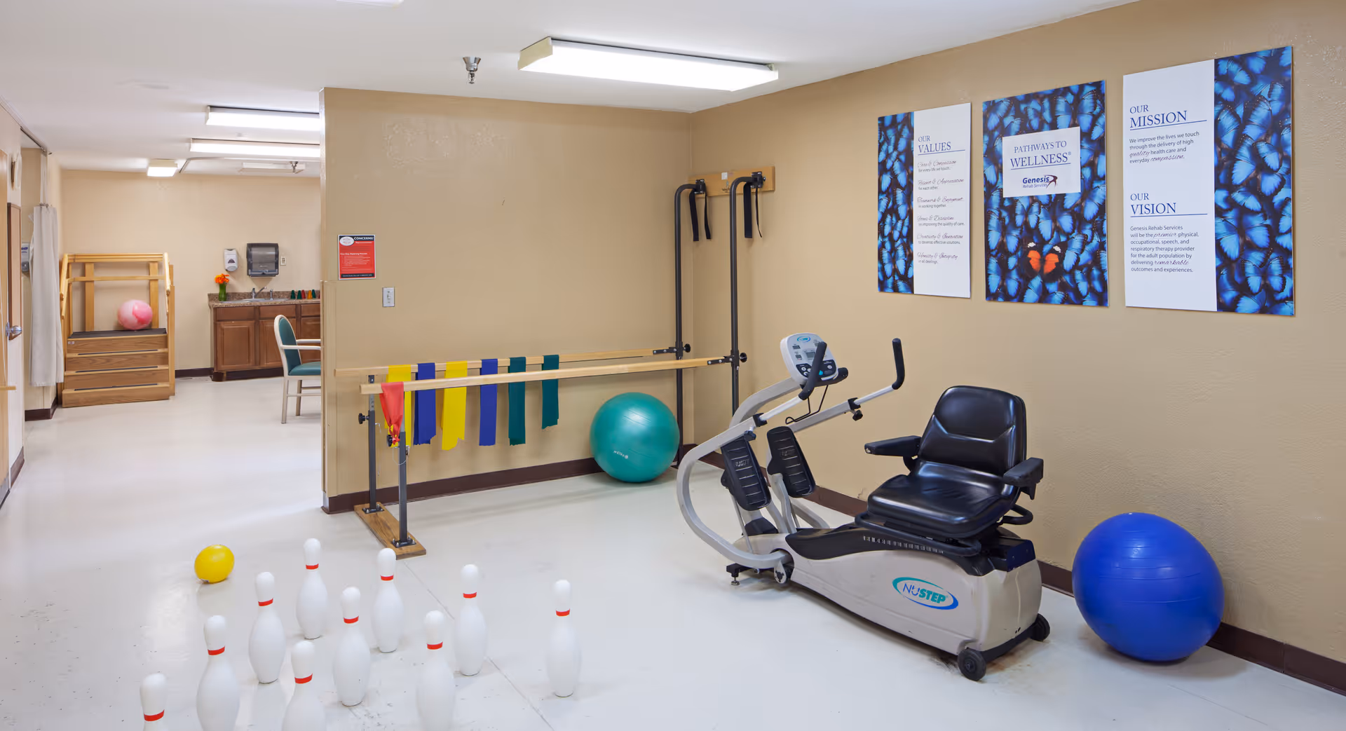A therapy or exercise room in a senior living facility with a recumbent exercise bike, colorful resistance bands hanging on a wooden rail, exercise balls, and a set of bowling pins arranged on the floor. The walls are beige with three posters displaying the facility's values, mission, and vision.