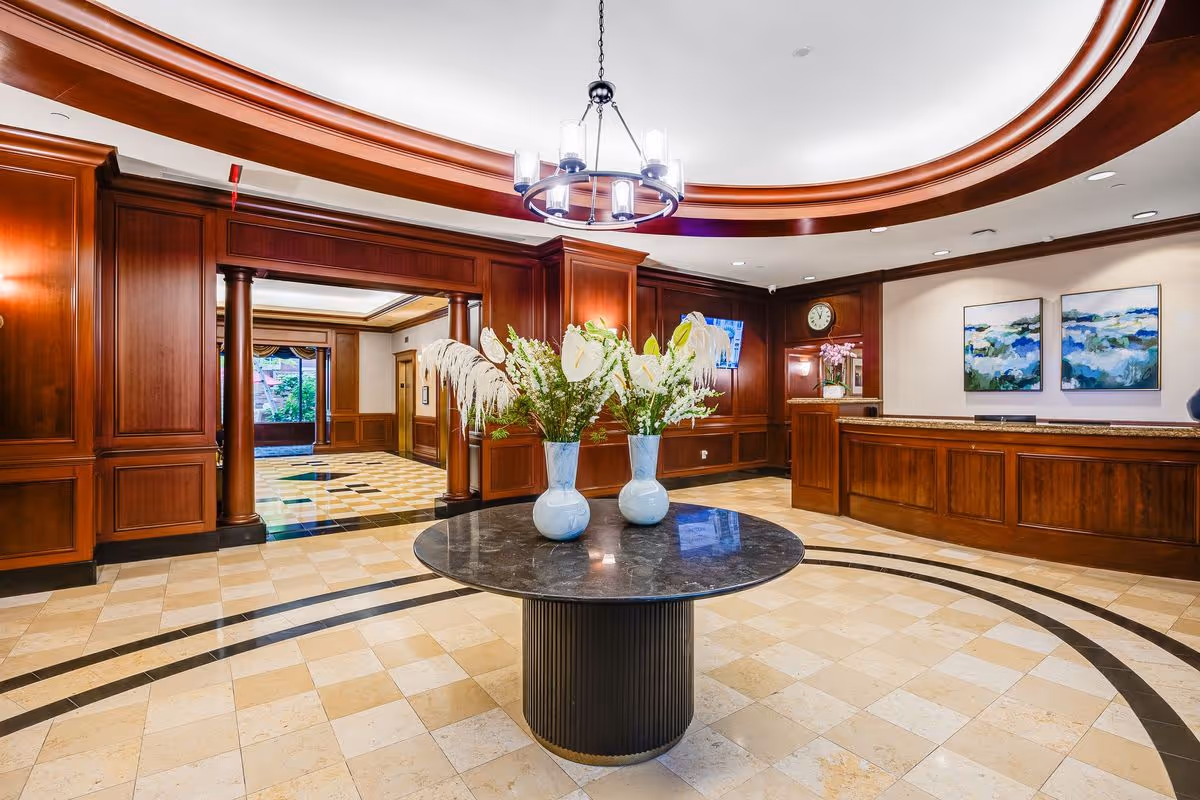 A spacious and elegant reception area with a round black marble table in the center holding two vases of white flowers. The room features polished beige tile flooring with dark inlaid patterns, rich wooden paneling on the walls, a chandelier hanging from a recessed ceiling with wood trim, and a wooden reception desk with a clock and two paintings on the wall behind it.