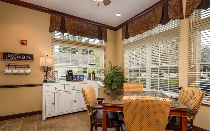 A cozy dining area with a wooden table surrounded by four cushioned chairs. The room has large windows with blinds and decorative valances, allowing natural light to fill the space. A white sideboard against the wall holds a coffee maker, cups, and other coffee supplies, with a small lamp and a sign above that reads 'coffee bar'. A green potted plant is placed in the corner near the windows.