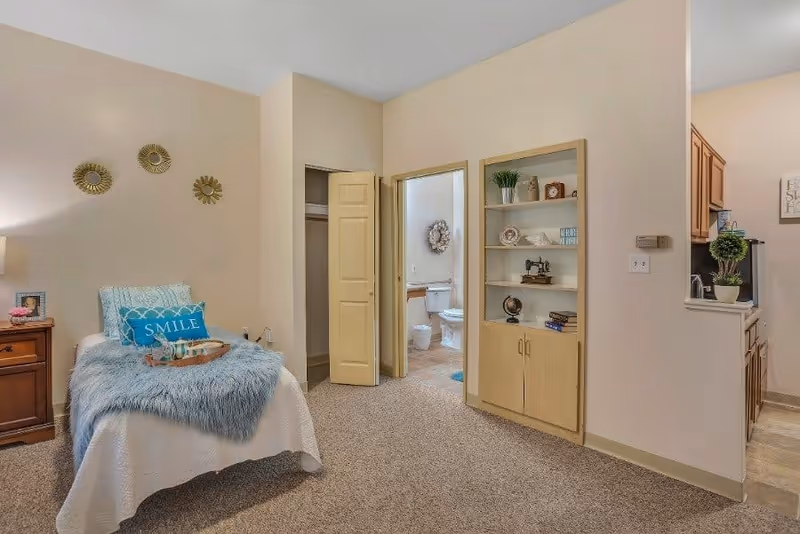 A cozy bedroom in an assisted living facility featuring a single bed with a blue throw blanket and a pillow that says 'SMILE'. There is a wooden nightstand with a lamp and picture frame beside the bed. The room has beige walls and carpeted floor. An open door reveals a bathroom with a toilet and a decorative wreath on the wall. A built-in shelving unit with various decorative items is visible next to the bathroom door. Part of a kitchen area with wooden cabinets and a small plant is seen on the right side.