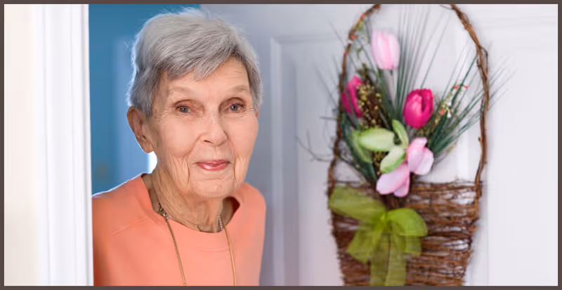 An elderly woman with short gray hair wearing a peach-colored top stands in a doorway, smiling gently. Next to her on the wall is a decorative basket with artificial flowers including pink and red tulips and green leaves.