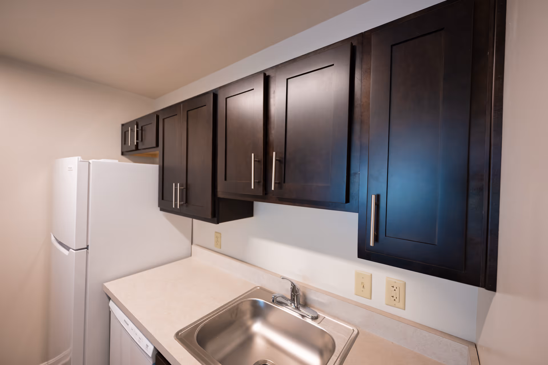 Small kitchenette with dark wood upper cabinets, a white refrigerator, light countertop and a stainless steel sink and faucet.