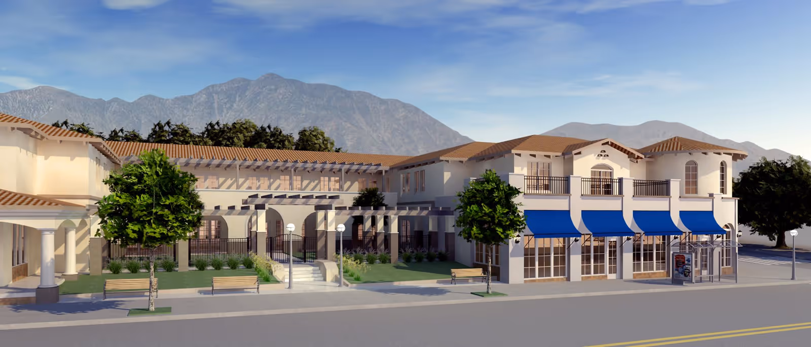 Exterior view of The Kensington Sierra Madre facility showing a two-story building with beige walls, red-tiled roofs, blue awnings over windows, benches, trees, and a mountain range in the background under a clear blue sky.