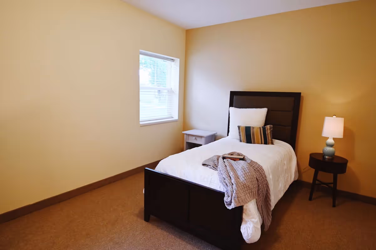 A simple bedroom with a single bed dressed in white bedding, a nightstand with a lamp, and a window on beige walls.