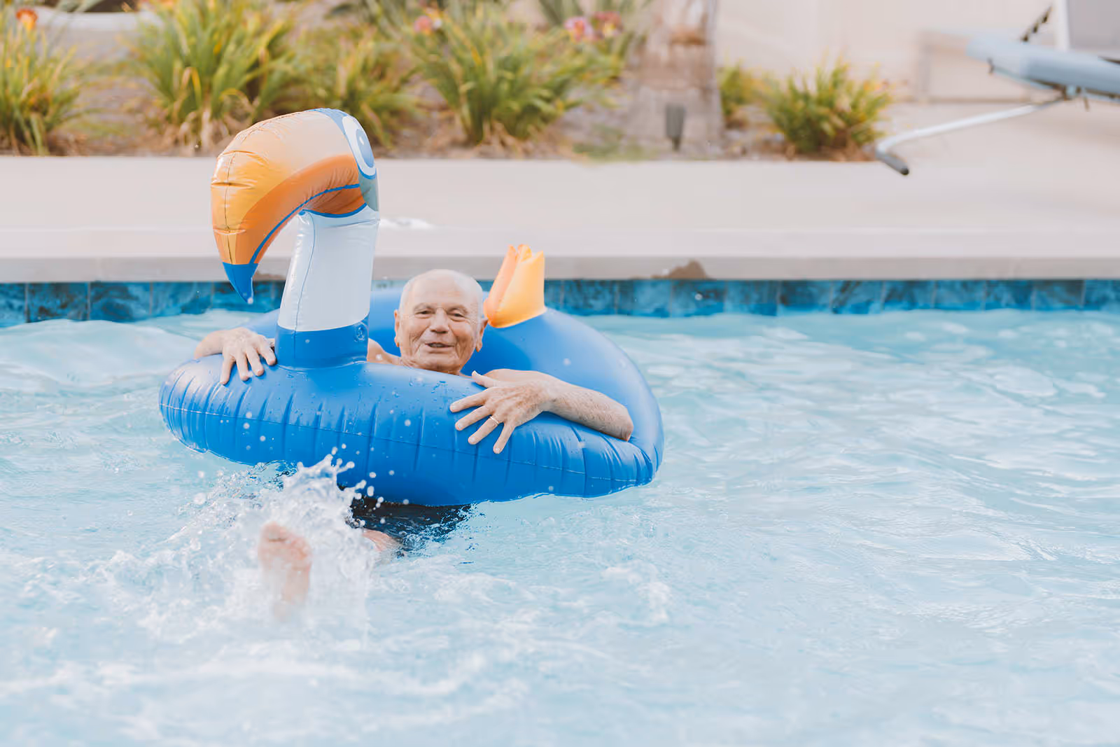 An elderly man enjoying himself in a swimming pool while holding onto a large blue inflatable float shaped like a bird with an orange beak.