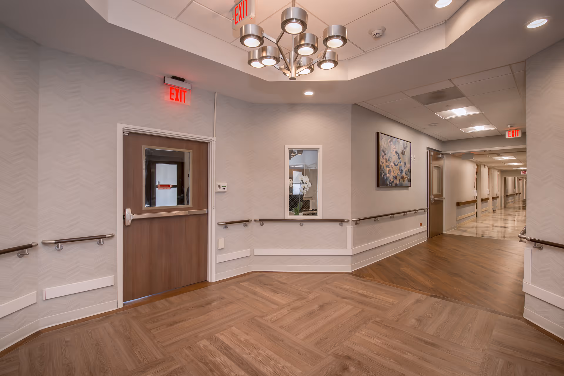 Interior view of a hallway in a senior living facility with wood-patterned flooring, beige walls with a subtle pattern, handrails along the walls, a wooden door with a window, multiple exit signs, a modern ceiling light fixture, and a framed artwork on the wall.