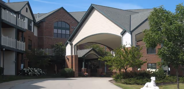 Exterior view of Meadowmere & Mitchell Manor Oak Creek showing a large covered entrance with a peaked roof, surrounded by brick and siding buildings, landscaped greenery, and a white dog statue on the right side near the driveway.