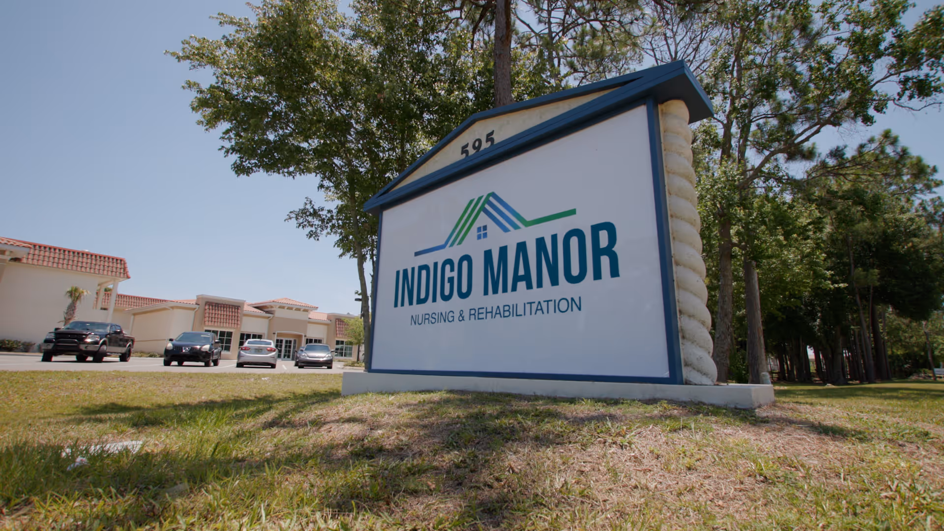 Outdoor view of a large sign for Indigo Manor Nursing & Rehabilitation with a building and parked cars in the background under a clear sky.