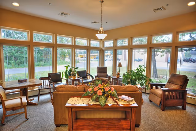 A bright and cozy living room with large windows letting in natural light. The room features a brown sofa, two armchairs, a wooden table with a floral centerpiece, several potted plants, and a small dining table with chairs near the windows. Outside, trees and parked vehicles are visible.