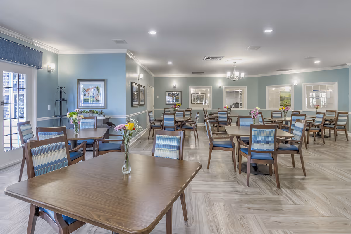 A bright and spacious dining room with multiple wooden tables and chairs arranged neatly. Each table has a small vase with colorful flowers. The walls are painted light blue with white trim, and there are several framed pictures and windows on the walls. The floor has a light wood pattern, and there is a chandelier hanging from the ceiling.
