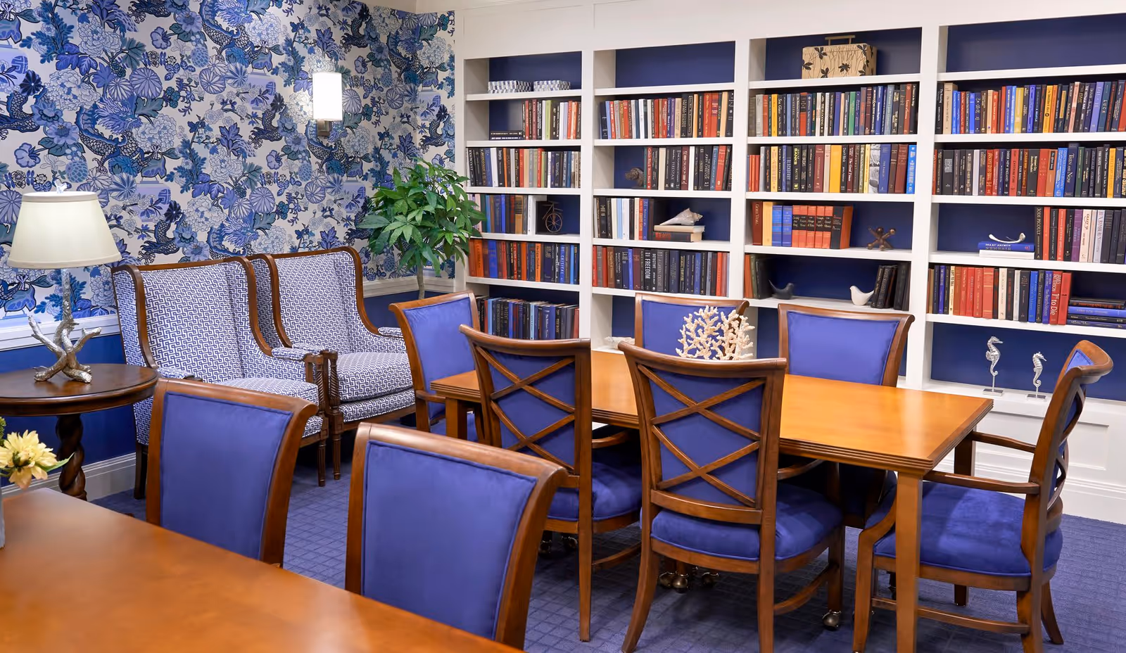 A cozy reading or meeting room with a wooden table surrounded by blue upholstered chairs. Behind the table is a large white bookshelf filled with books and decorative items. To the left, there are two patterned armchairs next to a small round wooden table with a lamp and a plant. The walls feature blue floral wallpaper and a wall sconce light.