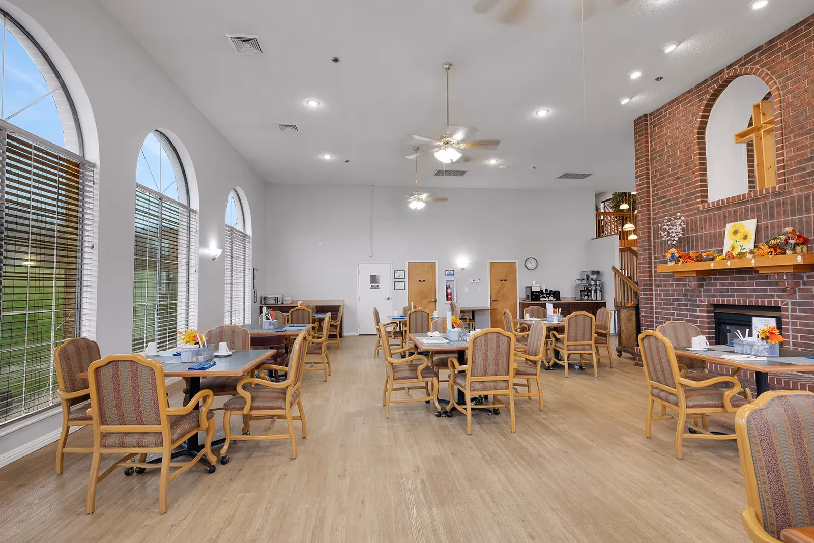 A spacious dining room with multiple tables and cushioned chairs arranged neatly. Large arched windows with blinds allow natural light to fill the room. The room features a brick fireplace decorated with flowers and artwork. Ceiling fans and recessed lighting are installed on the white ceiling. A coffee station and additional seating are visible in the background.